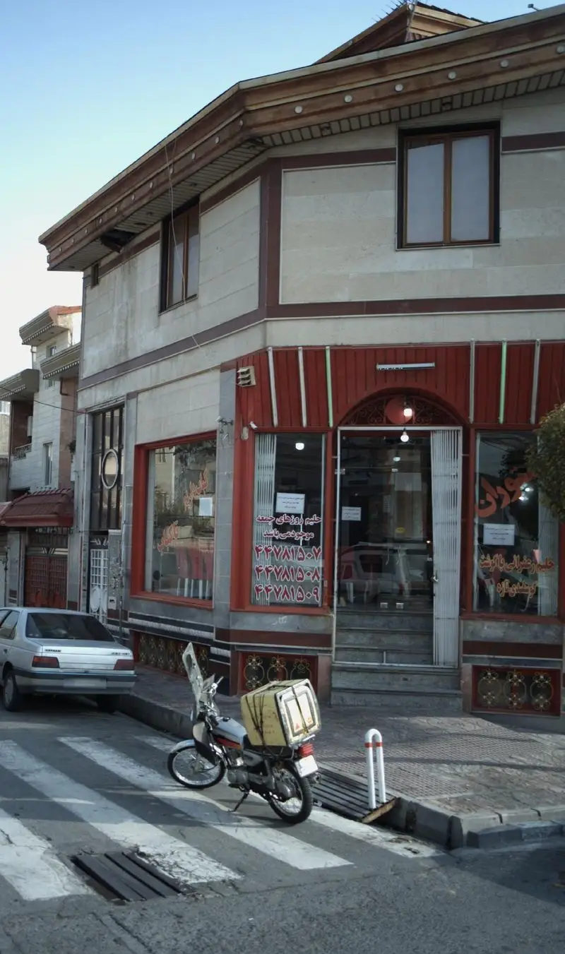 تصویر نمای بیرونی یک رستوران شلوغ در خیابانی پرتردد، با موتور سیکلت پیک برای تحویل غذای آماده و تابلو حلیم.