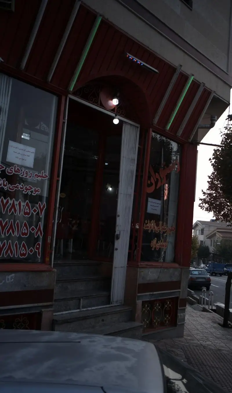 تصویری کامل از نمای بیرونی یک رستوران سنتی ایرانی در شهر، با درب ورودی باز، ویترین دارای شماره تلفن و اطلاعیه حلیم، و سقف چوبی قرمز.