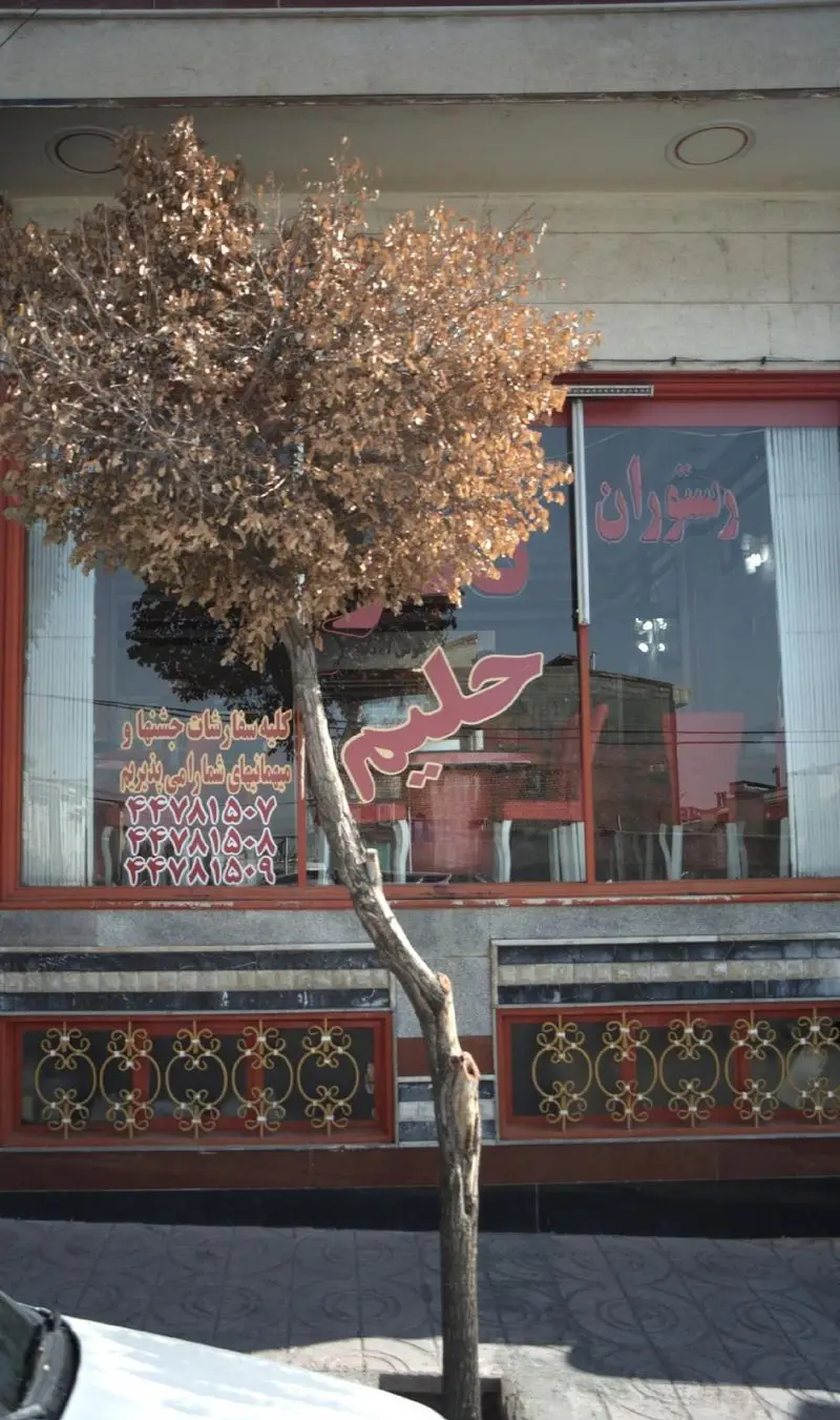 رستوران حلیم، پذیرای سفارشات جشن و میهمانی. نمای بیرونی رستوران با تابلوهای قرمز رنگ و شماره تلفن‌های تماس برای سفارش غذا.