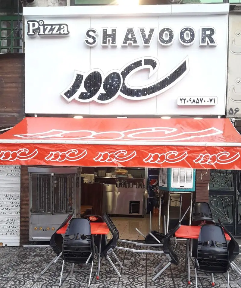تصویر نمای خارجی پیتزا شاوور با تابلوی درخشان فارسی و انگلیسی، میز و صندلی‌های روباز، رستوران فست‌فود آماده پذیرایی.