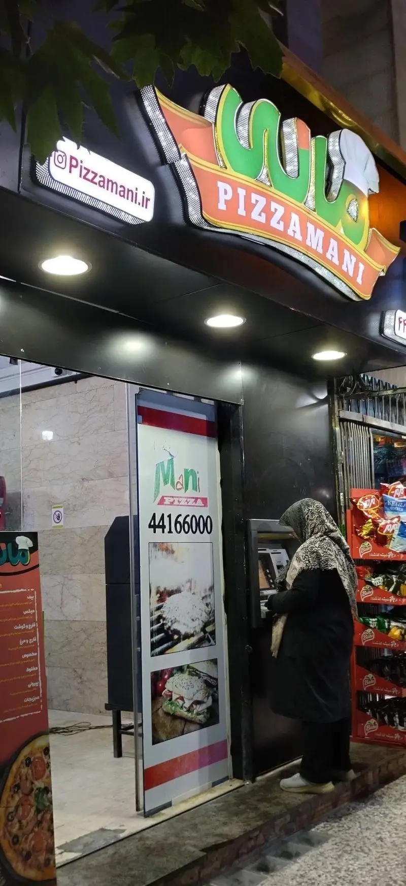 نمای شب فست فود پیتزا مانی با تابلوی روشن، شماره تلفن 44166000 و آدرس اینستاگرام Pizzamani.ir، نمایش پیتزا و برگر در منو.