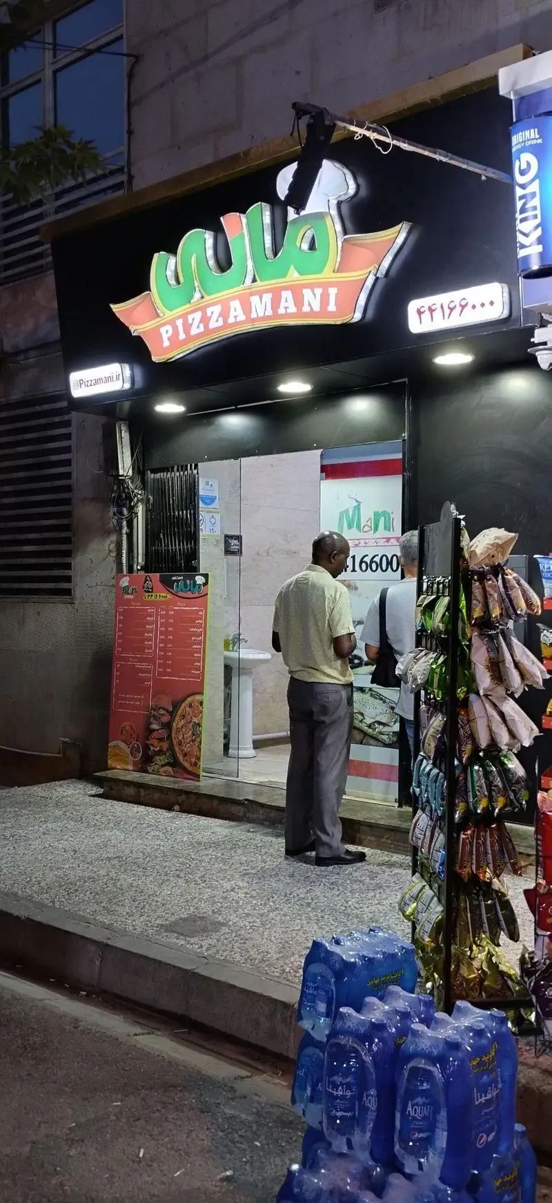 تصویر نمای بیرونی پیتزا مانی در شب، با تابلو، منو غذا، شماره تماس ۴۴۱۶۶۰۰ و محصولات فست فود. پیتزا، تنقلات و نوشیدنی.