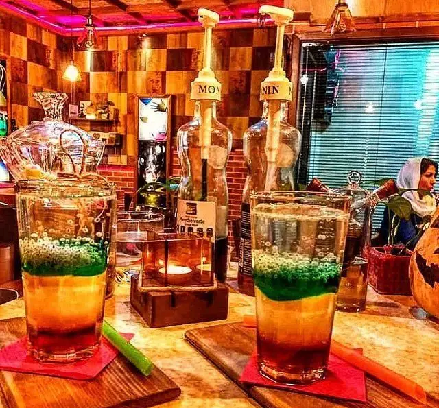 دو نوشیدنی لایه‌ای و رنگارنگ در محیط دلنشین کافه پندار با دکوراسیون چوبی و نورپردازی گرم، مکانی ایده‌آل برای استراحت و گپ‌وگفت.