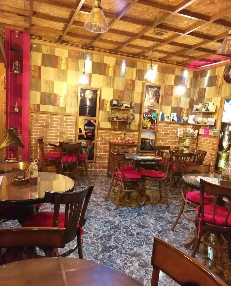 دکوراسیون داخلی کافه پندار، کافه‌ای دنج با میز و صندلی‌های چوبی و کوسن‌های قرمز، دیوارهای آجری و سقف چوبی، فضایی آرامش‌بخش و گرم.