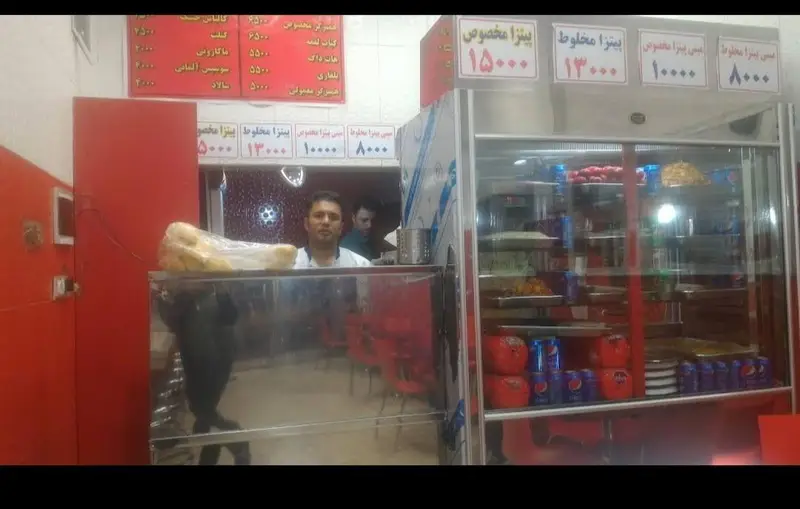نمایی از داخل رستوران فست فود اکبر بندری با منوی قیمت پیتزا و ساندویچ، ویترین پر از نوشیدنی‌ها و غذاهای آماده برای مشتریان.