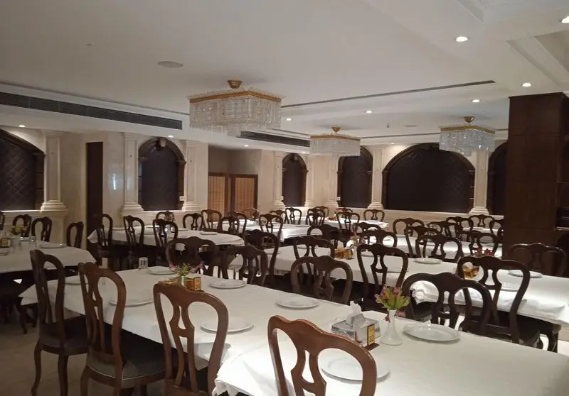 تصویری از فضای داخلی رستوران اقدسیه، نمایانگر محیطی بزرگ و شیک با میز و صندلی‌های چوبی کلاسیک و لوسترهای کریستالی زیبا برای پذیرایی.