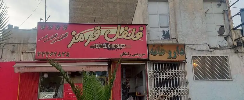 نمای بیرونی ساندویچی فلفل قرمز با تابلوی قرمز بزرگ، لوگوی فلفل و شماره تماس. نوشته سرویس رایگان و تابلوی داروخانه کناری.