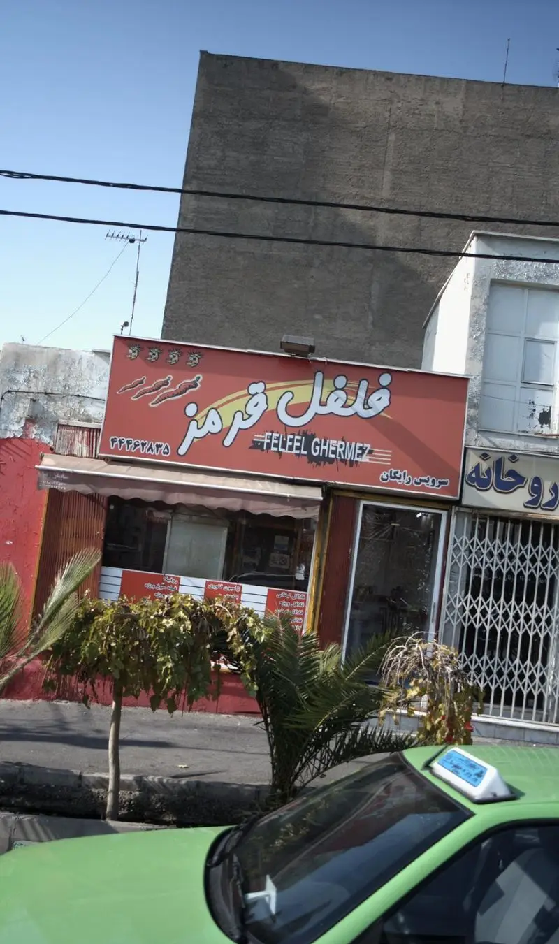 تصویر تابلوی فست فود فلفل قرمز (FELFEEL GHERMEZ) با سرویس رایگان و نماد فلفل چیلی در یک روز آفتابی، تاکسی سبز.