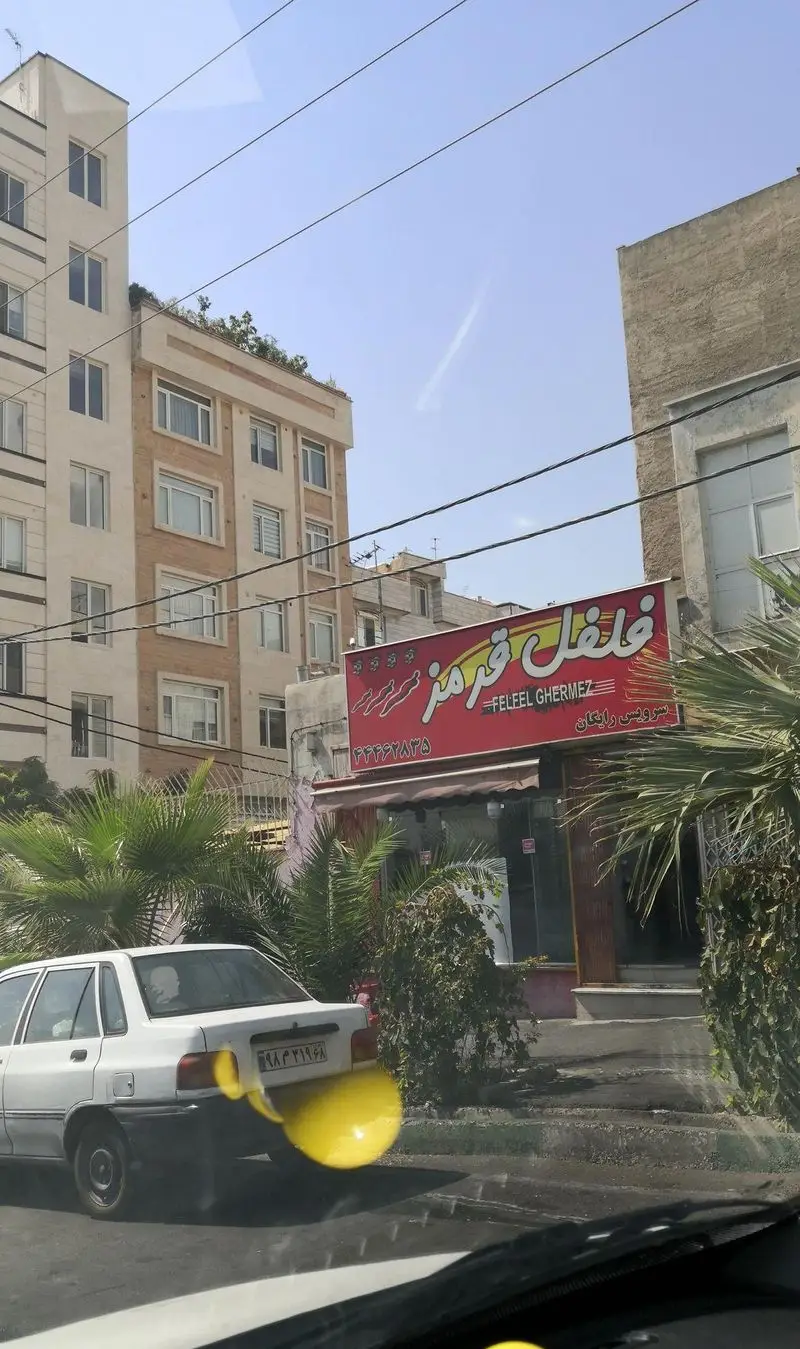 نمای بیرونی فست فود فلفل قرمز (Felfel Ghermez) با تابلوی قرمز، شامل شماره تماس و سرویس رایگان در روزی آفتابی، با خودرو و نخل.