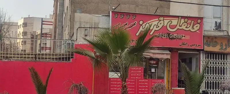 تصویر نمای بیرونی رستوران فلفل قرمز با رنگ قرمز جذاب، تابلوی بزرگ حاوی نام فارسی و انگلیسی، چند نخل در حیاط و سرویس رایگان. در کنار آن داروخانه دیده می‌شود.