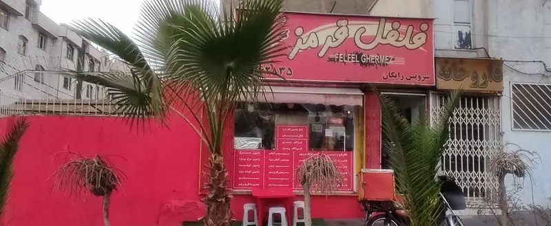 تصویر فروشگاه فلفل قرمز با نمای قرمز روشن، نخل‌های زیبا، میز و صندلی‌های فضای باز، موتور تحویل و تابلوی سرویس رایگان در کنار داروخانه.