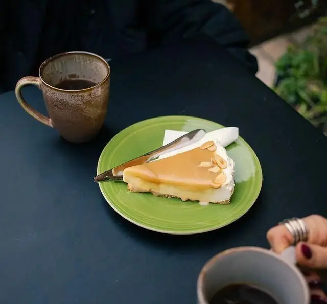 برش چیزکیک کارامل و بادام با قهوه تازه روی میز مشکی، در محیطی دنج و آرام از کافه رستوران دایموند. دسر و نوشیدنی عالی.