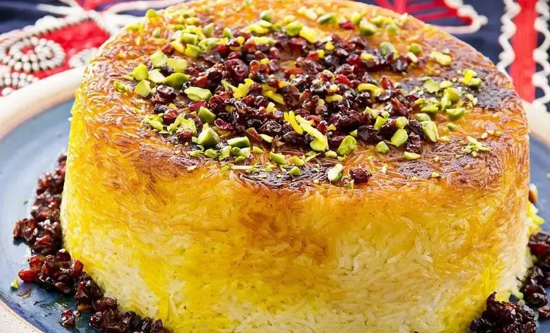 ته‌چین زعفرانی مجلسی با ته دیگ طلایی و خوش‌رنگ، تزئین شده با زرشک و خلال پسته تازه، یک غذای خاص از کترینگ رز برای مهمانی‌های شما.