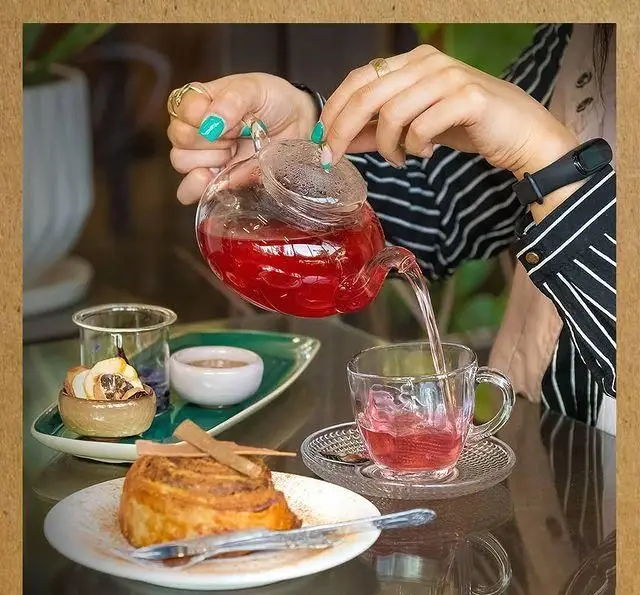 سرو گرم چای دمنوش قرمز از قوری شیشه‌ای به همراه رول دارچین خوشمزه در کافه دنج خانه فرهنگ و هنر سرهنگ.