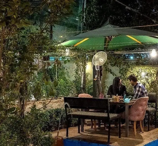 کافه فضای باز خانه فرهنگ و هنر سرهنگ در شب. محیطی دنج و سرسبز با چتر بزرگ، میز و صندلی‌ها برای پاتوقی دوستانه و گپ‌وگفت‌های صمیمی.