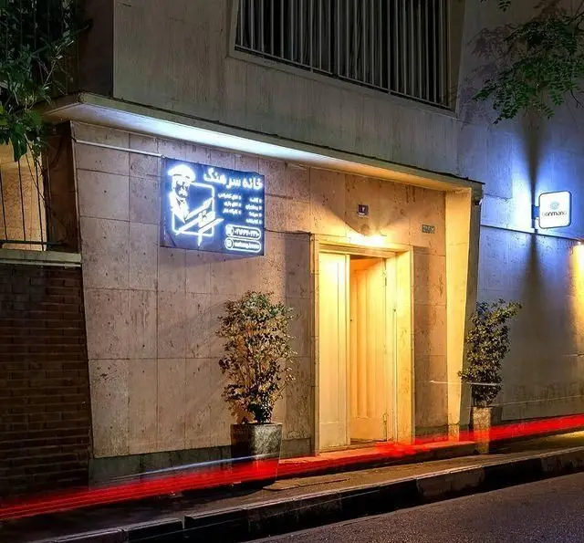 تصویر نمای بیرونی خانه فرهنگ و هنر سرهنگ در شب با تابلوی نئون روشن کسب و کار، ورودی نورانی و گلدان‌های تزئینی.