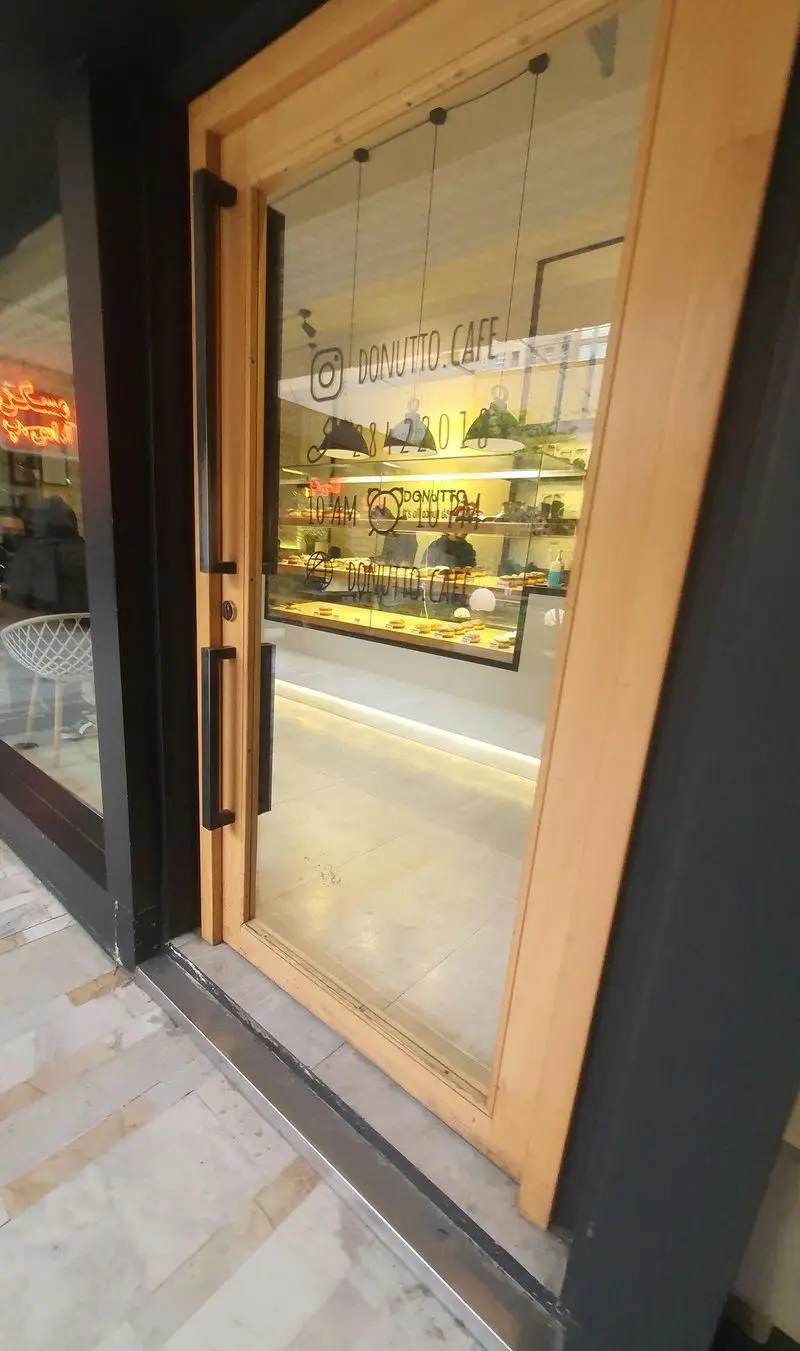 نمای بیرونی کافه دوناتو: ورودی شیک با درب چوبی و شیشه‌ای، ویترین پر از دونات و شیرینی‌های لذیذ، آدرس اینستاگرام DONUTTO.CAFE