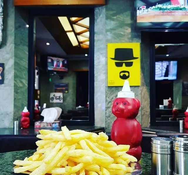 یک بشقاب سیب زمینی سرخ کرده تازه و طلایی در رستوران فست فود مستر برگر، با سس کچاپ خرسی و نماد معروف هایزنبرگ روی دیوار.