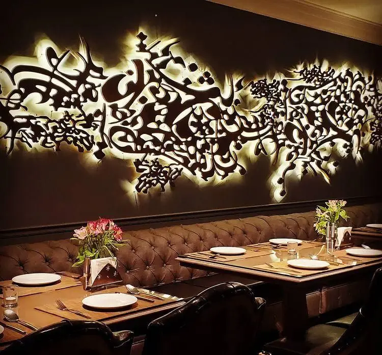 فضای داخلی لوکس کافه رستوران هرمان با دکوراسیون چشم‌نواز، خطاطی نورانی زیبا روی دیوار و میزهای چیدمان شده برای سرو غذا و نوشیدنی.