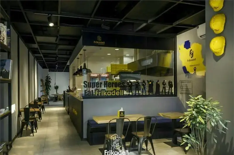 تصویری از فضای داخلی فست فود فریکادل، با دکوراسیون مدرن و تم سوپرهیرویی، آشپزخانه شیشه‌ای و میز و صندلی‌های راحت آماده برای پذیرایی.