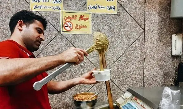 مردی آشپز در آش و حلیم پزی حاج علی معنوی و پسران در حال سرو حلیم داغ و خوشمزه برای مشتریان وفادار.