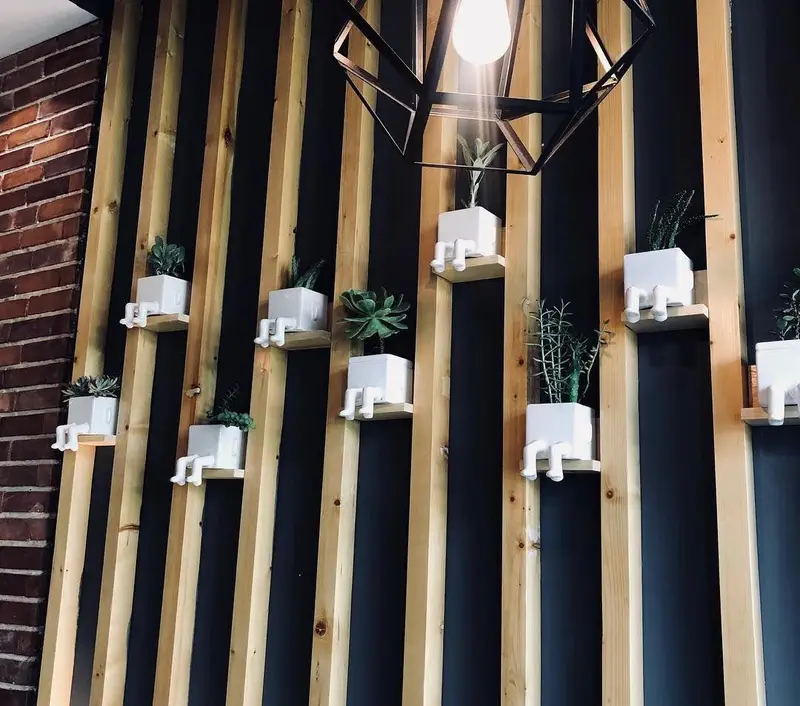 دکوراسیون داخلی پیتزا تنوری پاروت: دیوار چوبی عمودی با گلدان‌های فانتزی سفید نشسته و گیاهان سبز متنوع روی شلف‌های چوبی، فضایی دنج و جذاب.