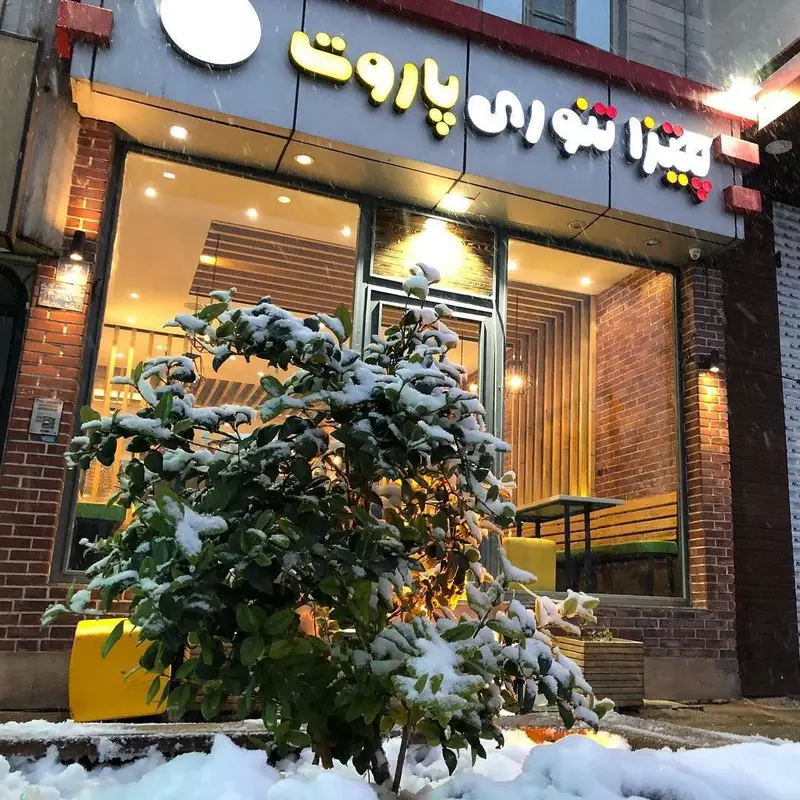 نمای بیرونی رستوران پیتزا تنوری پاروت در یک روز برفی زمستانی، با برف نشسته بر روی بوته جلوی مغازه و تابلوی درخشان.