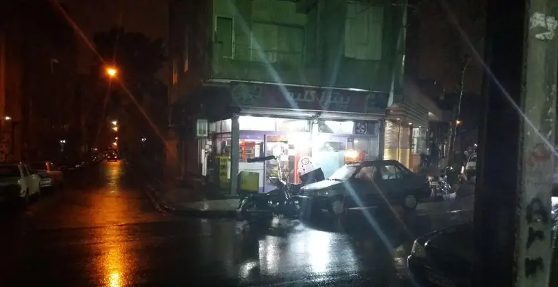 نمای شبانه پیتزا کلبه در خیابانی خیس. بازتاب نورهای شهری و مغازه بر آسفالت. یک ماشین و موتور سیکلت افتاده جلوی مغازه.