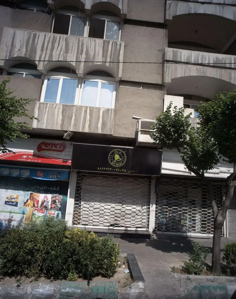 نمای بیرونی ساختمان پیتزا کلبه در محیط شهری، شامل مغازه‌های بل برگر و تکدانه با کرکره‌های بسته، درختان و پیاده‌رو.