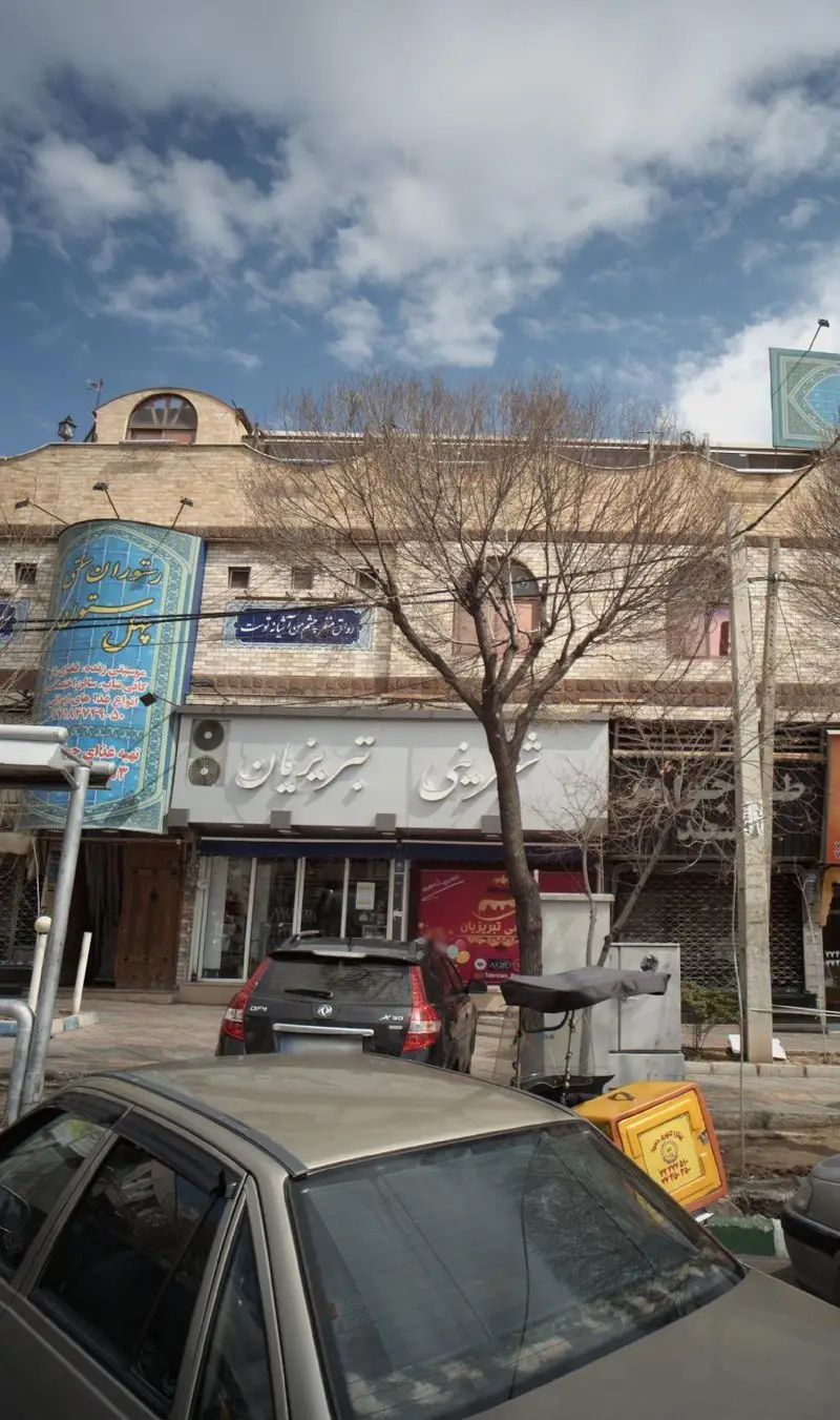 نمای بیرونی شیک و جذاب شیرینی تبریزیان در خیابان، با تابلوی بزرگ و واضح قنادی و ورودی فروشگاه، در کنار خودروها زیر آسمان ابری.