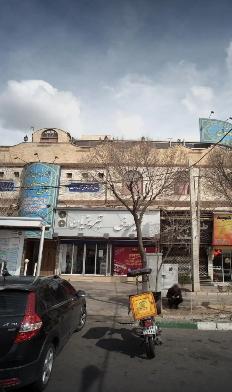 شیرینی تبریزیان: نمای بیرونی این فروشگاه شیرینی و قنادی در یک روز ابری با خودرو و موتورسیکلت پیک و مردم در خیابان.