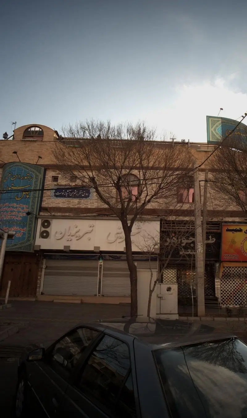 نمای بیرونی فروشگاه شیرینی تبریزیان، با تابلوی سفید بزرگ و درختان بدون برگ در خیابانی شلوغ در فصل پاییز یا زمستان.