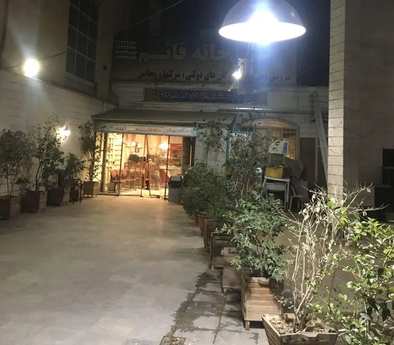 ورودی روشن تهیه غذای قائم در شب، کترینگ مجالس و شرکت‌ها با فضای دلنشین و گلدان‌های کنار راهرو.