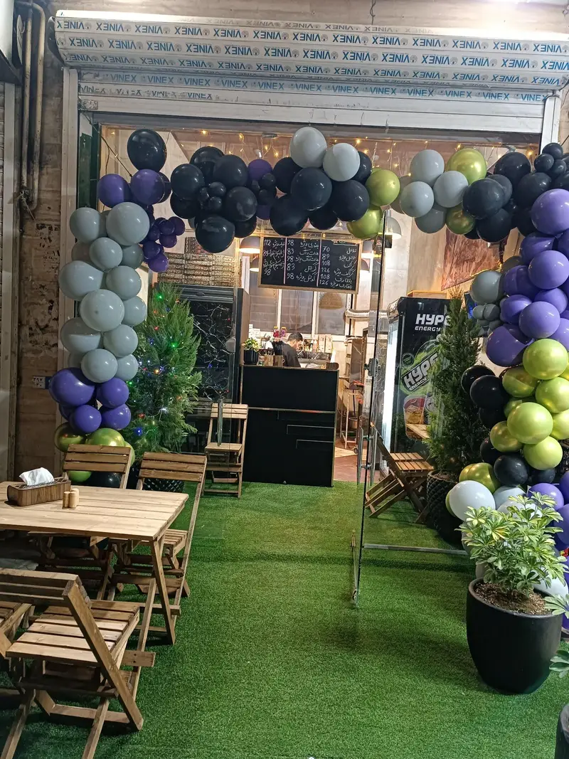 نمای ورودی فست فود وودی با طاق بادکنکی سیاه، سفید، بنفش و طلایی، فضای سبز بیرون بر، میز و صندلی چوبی و منوی روی تخته