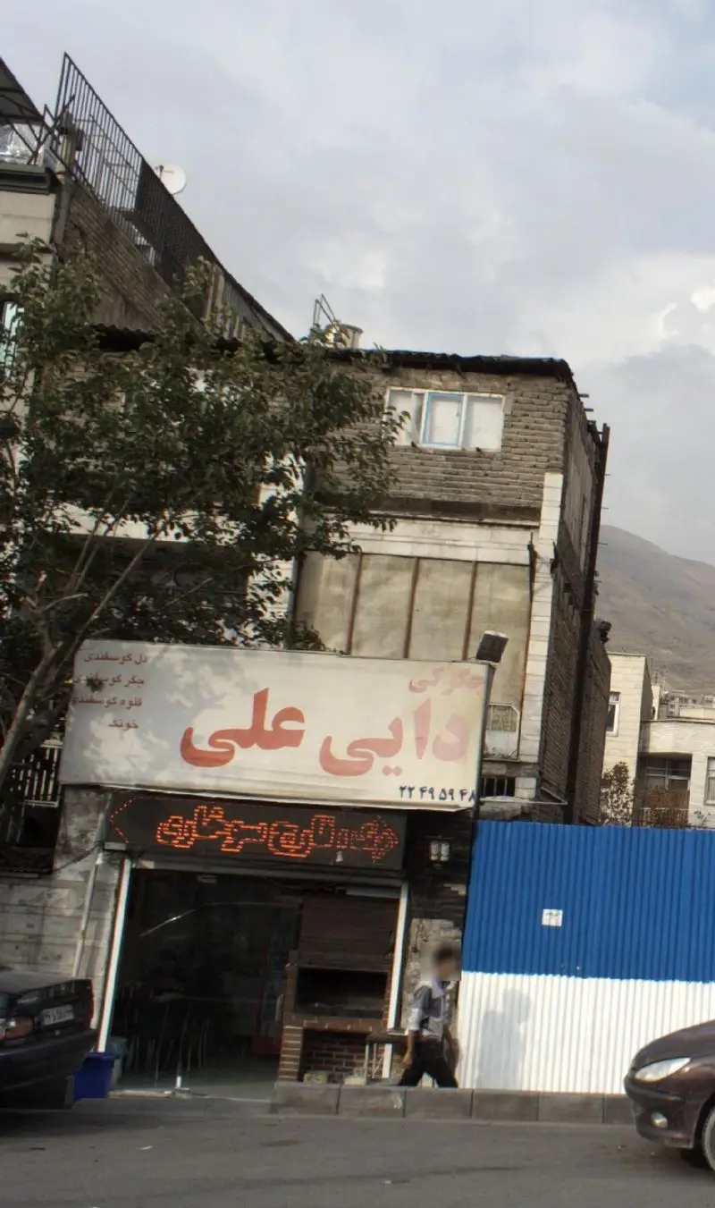 جگرکی دایی علی: نمای بیرونی مغازه عرضه کننده کباب جگر، دل و قلوه گوسفندی با تابلوی فارسی، درخت و کوه در پس زمینه، فضایی دلنشین.