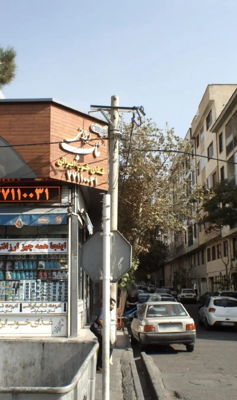 آشپزخانه ناب ایرانی، رستورانی با غذای خوب ایرانی در خیابانی شلوغ، شامل تابلوی فارسی، شماره تلفن ۷۱۱۰۰۳۱ و شعار 'اینجا همه چیز ارزان است'.