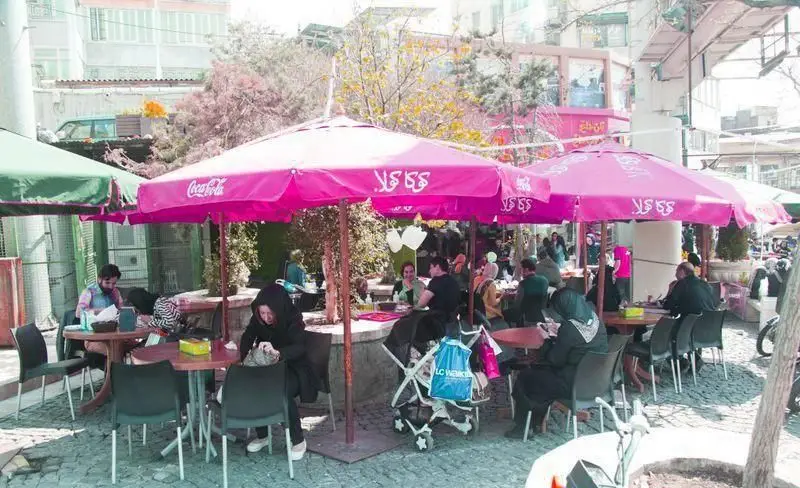مشتریان در فضای باز دلنشین و سرسبز رستوران توچال کباب، زیر چترهای صورتی کوکاکولا، از طعم لذیذ غذا و نوشیدنی‌های تازه لذت می‌برند.