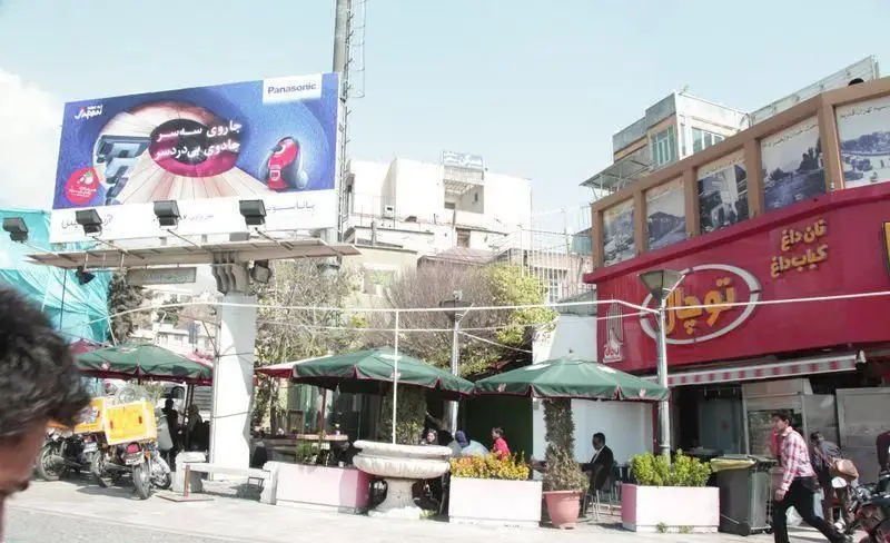 تصویر رستوران توچال کباب، ارائه دهنده کباب داغ و نان داغ، با میز و صندلی‌های فضای باز زیر چترهای سبز در خیابان شلوغ.