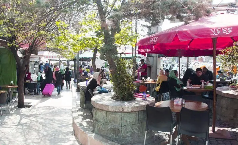 مشتریان در فضای باز رستوران توچال کباب زیر سایه درختان و چترهای قرمز مشغول غذا خوردن هستند، محیطی دلنشین و سرزنده برای تجربه کباب.