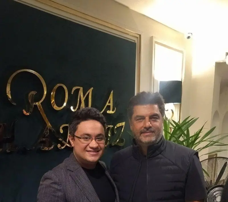 تصویری از دو مهمان خندان در شاندیز روما، رستوران معروف با تابلوی طلایی ROMA در پس‌زمینه. فضایی لوکس برای تجربه غذایی.