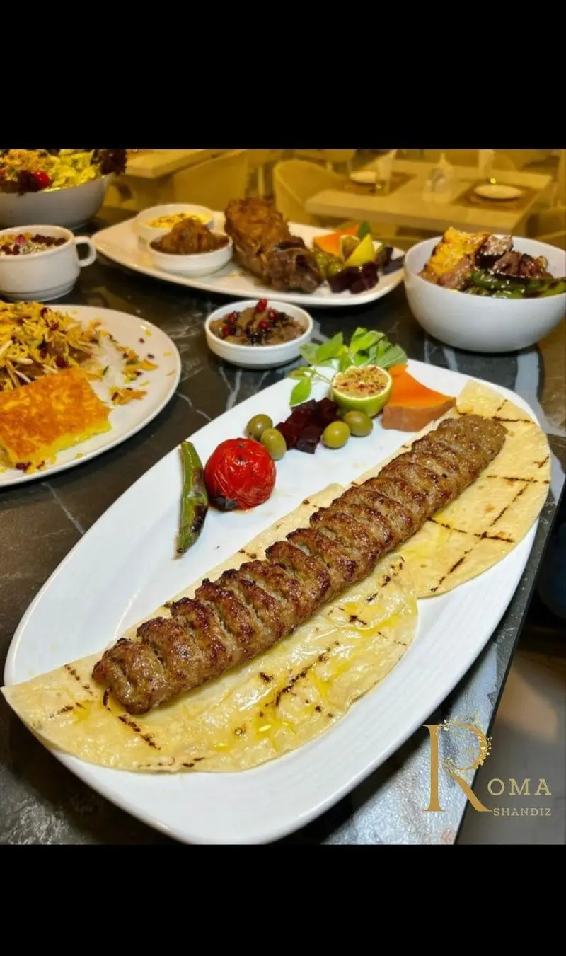 کباب کوبیده ممتاز شاندیز روما، با نان داغ، گوجه، فلفل و زیتون، در کنار میز پر از برنج زعفرانی و انواع غذاهای خوش طعم ایرانی.