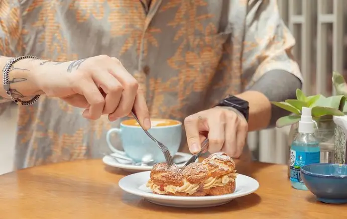 لذت بردن از دسر و قهوه در کافه بالا. مشتری در حال برش شیرینی خامه‌ای با قهوه لاته داغ. یک تجربه خوشمزه در فضایی دلپذیر.