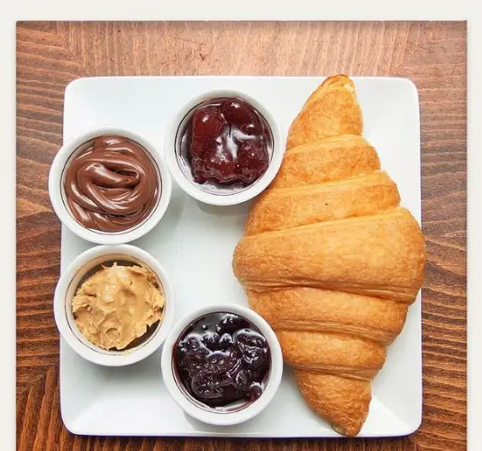 یک کرواسان تازه و طلایی از کافه ژی، همراه با مرباهای توت فرنگی و بری، نوتلا و کره بادام زمینی برای صبحانه یا عصرانه لذیذ.