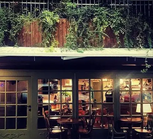 نمای دنج و دلنشین کافه ژی در شب. نور گرم داخلی، میز و صندلی‌های چوبی از پشت پنجره‌های بزرگ و فضای سبز زیبا بالای بالکن.