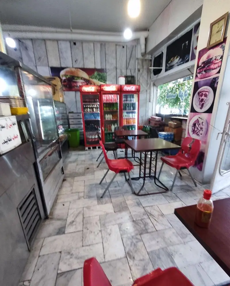 فست فود چلچله: فضای داخلی دنج با میز و صندلی‌های قرمز، یخچال‌های نوشیدنی کوکاکولا، پیشخوان غذا، و پوسترهای بزرگ برگر و پیتزا.