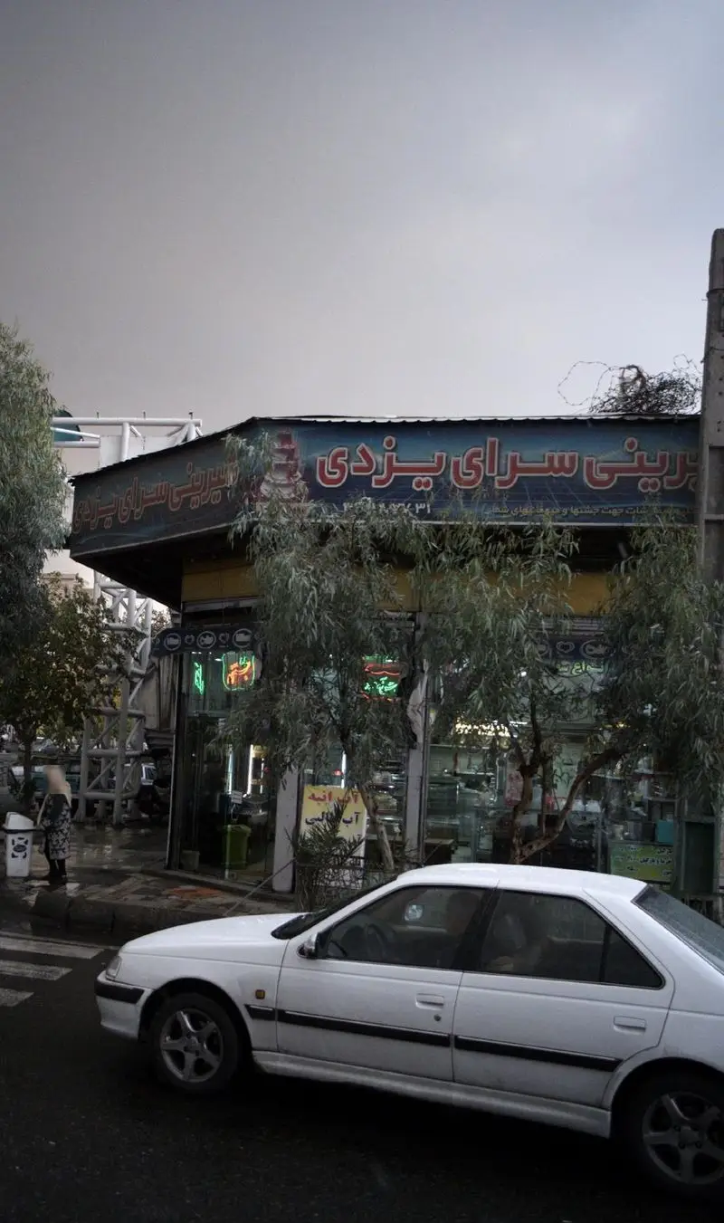 تصویر شیرینی سرای یزدی، قنادی معروف با تابلوی آبی و قرمز رنگ، درختان سبز جلوی مغازه و خودروی سفید در خیابان.