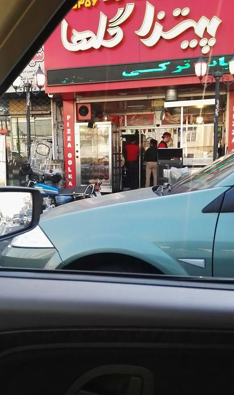 نمای بیرونی رستوران پیتزا گلها در یک روز شهری، با تابلوی بزرگ و روشن، کارکنان مشغول کار و موتورسیکلت در جلوی فروشگاه.