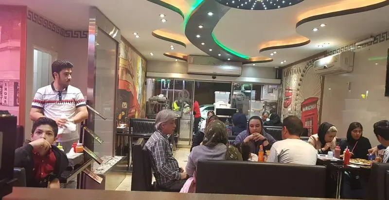 پیتزا گلها: فضای داخلی شیک و مدرن رستوران با سقف‌های نورانی، مشتریان مشغول صرف پیتزا و غذا در کنار خانواده و دوستان.