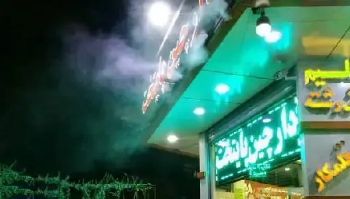 تصویر شب آشکده دارچین با بخار متصاعد از بالای سقف، تابلوی نئون سبز و روشنایی محیط، نشان دهنده یک آش فروشی پرطرفدار.