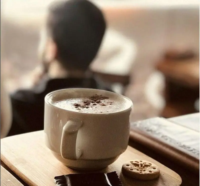 یک فنجان کاپوچینو یا لاته خوش عطر کافه گندم با پودر کاکائو روی میز چوبی، همراه با بیسکویت و شکلات برای لحظاتی آرامش‌بخش.